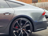 A sleek silver car partially visible, parked on a street. Its prominent black alloy wheel displays a shiny finish. Red brake calipers peek through. Background features a brick wall and wooden fence.
