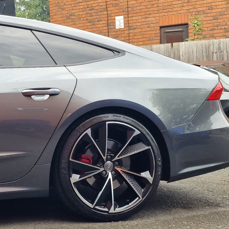 A sleek silver car with distinct alloy wheels and visible logo is parked on a street. It is set against a brick wall and wooden fence background.