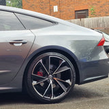 Sleek silver car; parked on asphalt with a prominent glossy black and silver alloy wheel; against a backdrop of brick building and wooden fence.