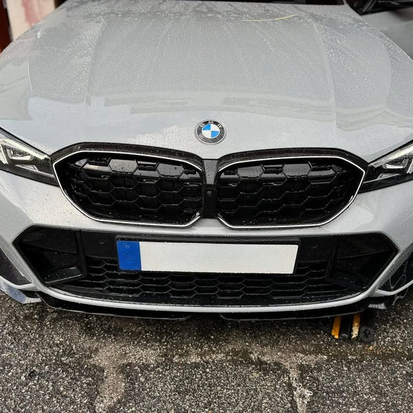A white BMW car front with a distinctive grille sits on wet pavement, flanked by a brick wall and an open door, displaying a European-style license plate.