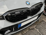 A silver BMW car front displays a prominent grille with a logo caught in light rain, parked on dark, wet asphalt.