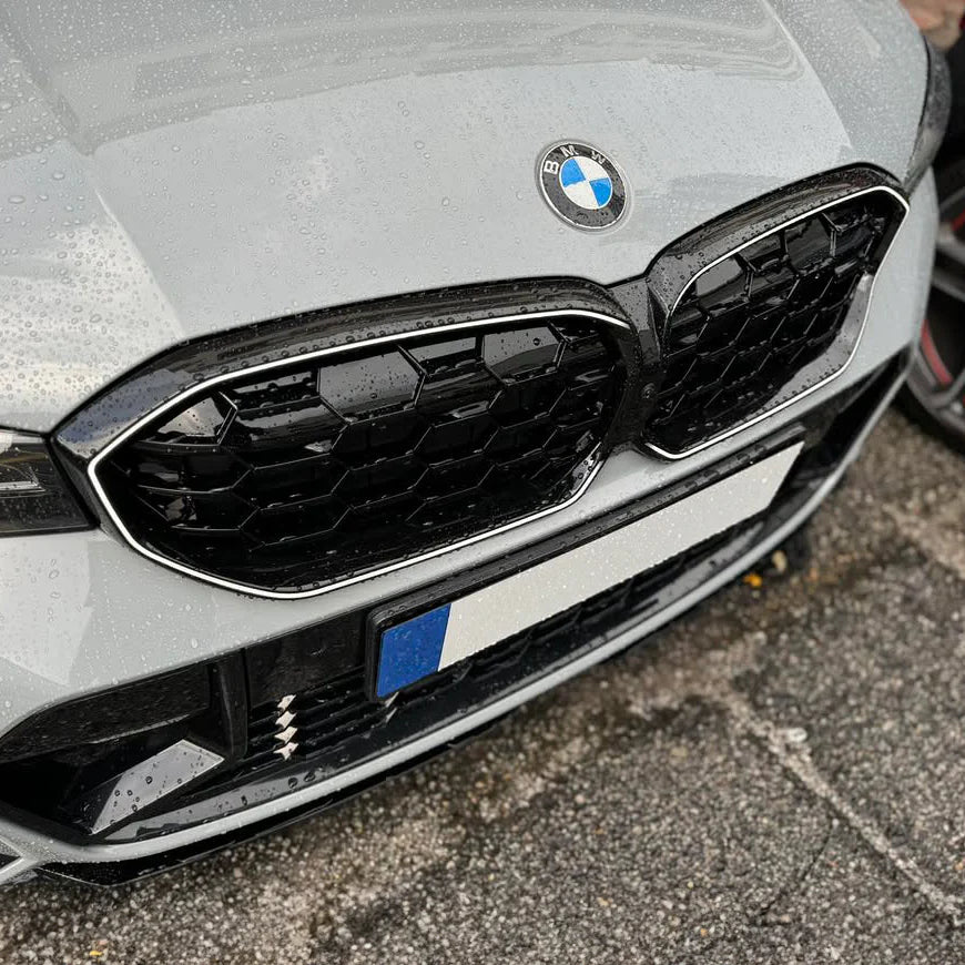 A silver BMW car front displays a prominent grille with a logo caught in light rain, parked on dark, wet asphalt.