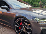 A sleek gray car with distinctive black alloy wheels and red brake calipers is parked on a brick driveway. A house and greenery form the backdrop.
