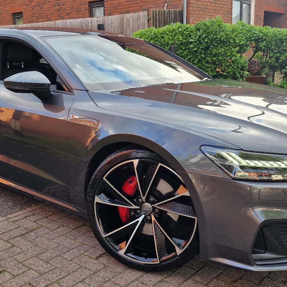 A sleek gray car with distinctive black alloy wheels and red brake calipers is parked on a brick driveway. A house and greenery form the backdrop.