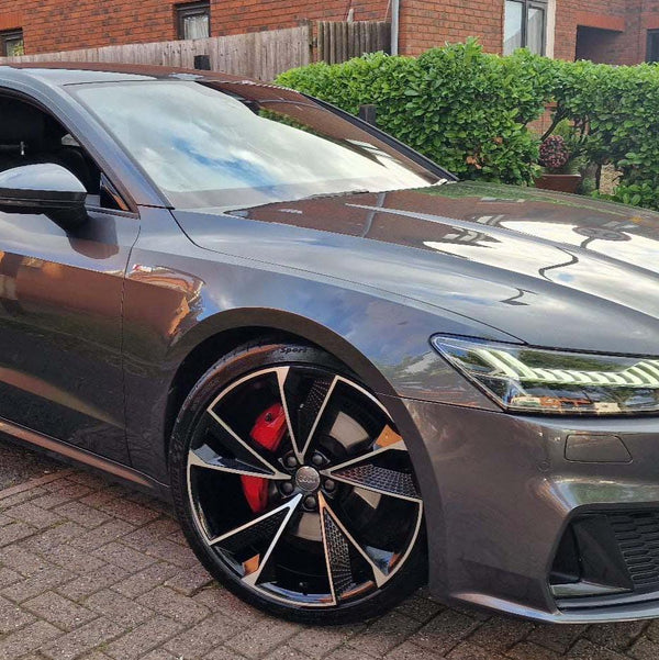 A sleek gray car parked on a paved driveway, showcasing shiny black alloy wheels and illuminated headlights, with a brick house and trimmed hedges in the background.