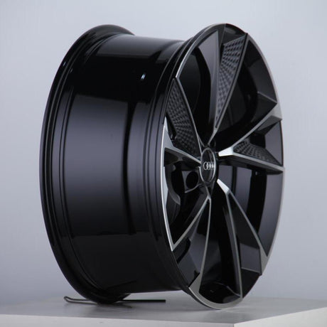 A glossy, black alloy car wheel featuring a complex, geometric design and a recognizable emblem at the center, rests on a plain, light-colored surface against a neutral backdrop.