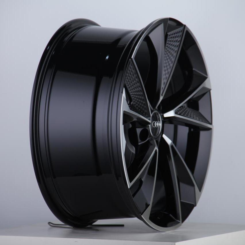A black car wheel rim with five angular spokes is displayed upright on a white surface; the center features a circular logo with interconnected rings.