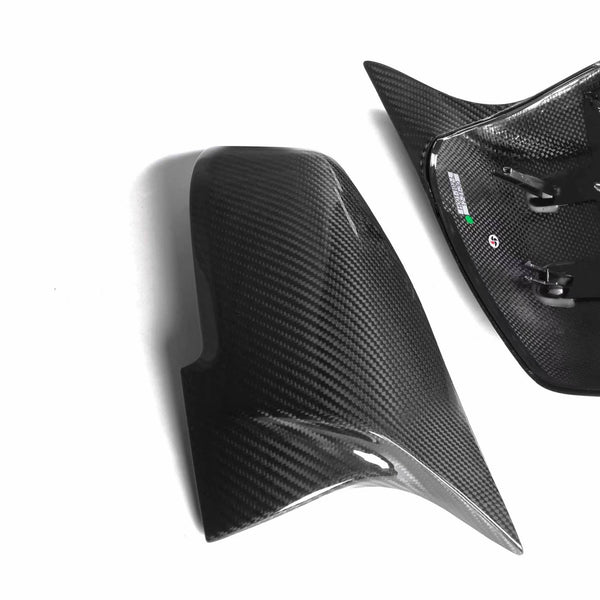 Carbon fiber mirror covers, one with mounting clips, rest on a white background. The shiny surface reflects light, highlighting the weave pattern for a sleek automotive appearance.
