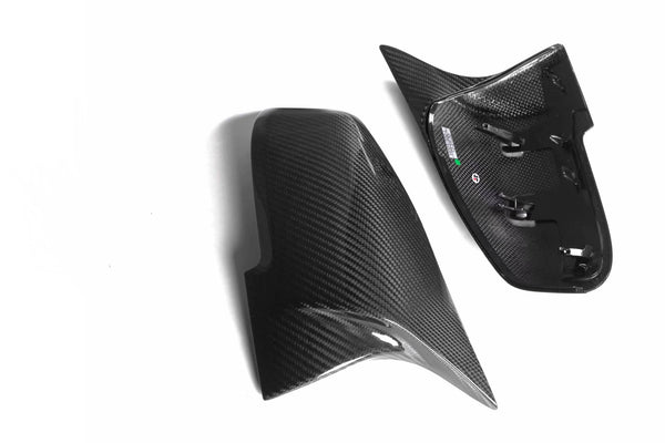 Carbon fiber mirror covers, one with mounting clips, rest on a white background. The shiny surface reflects light, highlighting the weave pattern for a sleek automotive appearance.