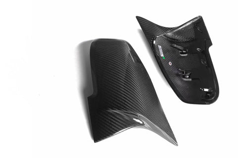Carbon fiber mirror covers, one with mounting clips, rest on a white background. The shiny surface reflects light, highlighting the weave pattern for a sleek automotive appearance.