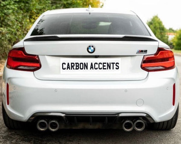 A white BMW M2 is parked on a road, featuring a "CARBON ACCENTS" license plate. Surrounded by greenery, dual exhausts are visible beneath the rear bumper.