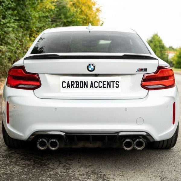 A white BMW M2 is parked on a road, featuring a "CARBON ACCENTS" license plate. Surrounded by greenery, dual exhausts are visible beneath the rear bumper.