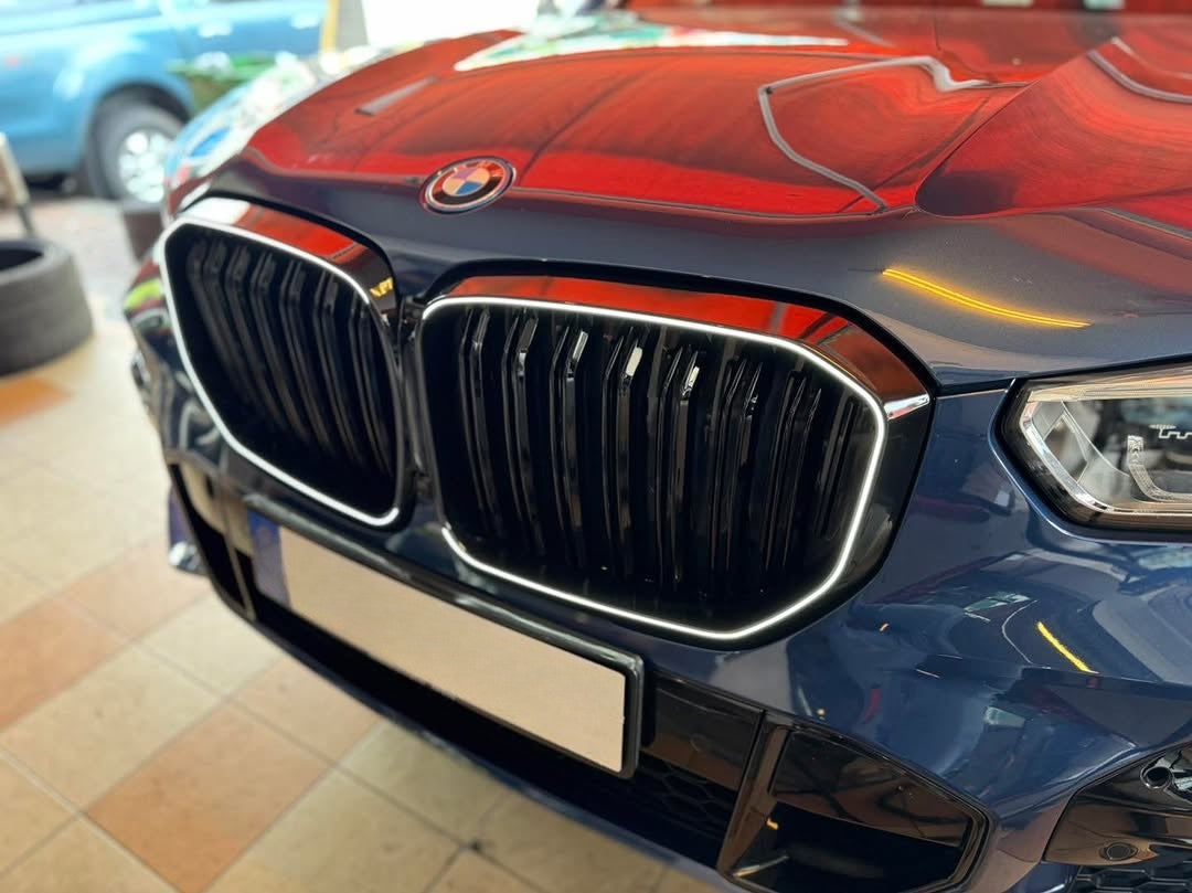 The front of a blue BMW features a prominent grille and sleek headlights in a tiled, indoor area. The car's hood reflects a vivid red hue from overhead lighting.