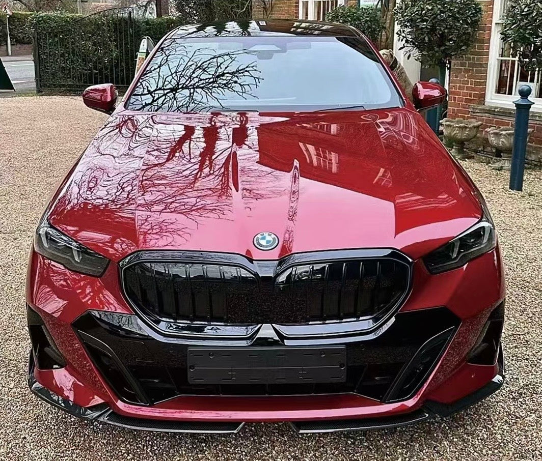 A red BMW car is parked on a gravel driveway, reflecting tree branches on its polished surface, with a brick building and greenery surrounding it.
