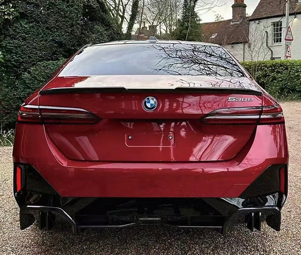 A red BMW 5-series car is parked on a gravel driveway. The setting includes green hedges and trees, with a brick house in the background.