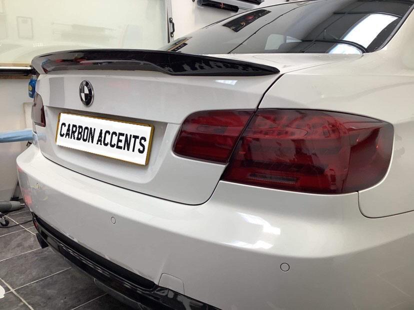 A white car displays a carbon fiber spoiler, parked in a garage. The rear license plate reads "CARBON ACCENTS."