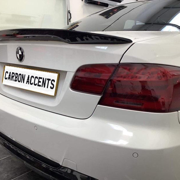 A white car displays a carbon fiber spoiler, parked in a garage. The rear license plate reads "CARBON ACCENTS."