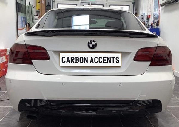 A white BMW is parked indoors, displaying a license plate reading "CARBON ACCENTS." The garage setting includes tiled flooring and automotive tools arranged on racks.
