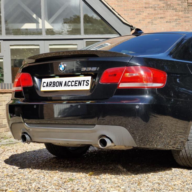 A black BMW 335i is parked on a gravel driveway, with a license plate reading "CARBON ACCENTS," against a background of a brick building with large windows.