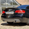 A black BMW 335i is parked on a gravel driveway, with a license plate reading "CARBON ACCENTS," against a background of a brick building with large windows.