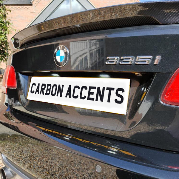 A black BMW 335i, parked near a brick wall, displays a license plate reading "CARBON ACCENTS" beneath a carbon fiber rear spoiler.