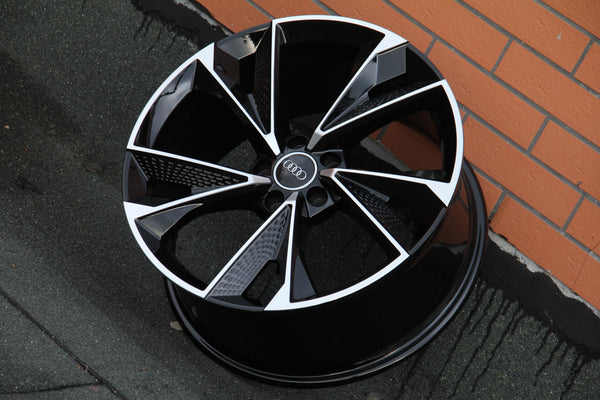 A glossy black and silver alloy wheel lies against an orange brick wall. The center has an Audi logo, surrounded by angular spokes. The ground is dark and textured.