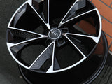 A black and silver Audi car wheel lies on a gray pavement next to an orange brick wall, featuring a glossy finish and intricate geometric spoke design.
