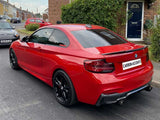 A red BMW car is parked on a residential street, showcasing black wheels and tinted windows. Nearby are other parked cars and brick houses surrounded by greenery. Text: 