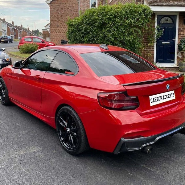 A red BMW car is parked on a residential street, showcasing black wheels and tinted windows. Nearby are other parked cars and brick houses surrounded by greenery. Text: "CARBON ACCENTS."