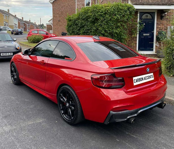 A red BMW car is parked on a residential street, showcasing black wheels and tinted windows. Nearby are other parked cars and brick houses surrounded by greenery. Text: "CARBON ACCENTS."