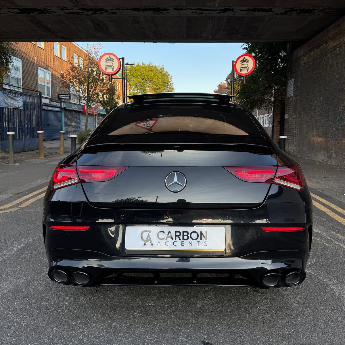 A black Mercedes car is parked under a bridge with two road signs indicating no entry for cars and motorcycles. License plate reads "CARBON ACCENTS." Urban street with buildings nearby.