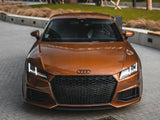 A bronze Audi sports car is parked on a brick-paved driveway surrounded by landscaped grass and modern buildings. The car's headlights are on, and its grille is prominently visible.