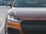 A bronze Audi sports car sits parked, its sleek front grille and headlights visible. It rests on a concrete pavement with metal barriers and a sunset sky in the background.