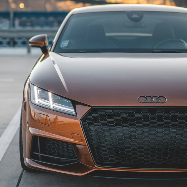 A bronze Audi sports car sits parked, its sleek front grille and headlights visible. It rests on a concrete pavement with metal barriers and a sunset sky in the background.