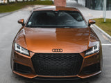 A metallic bronze Audi sports car is parked on a street near modern buildings and a grassy area. Its sleek design features angular headlights and a prominent grille with the Audi logo.