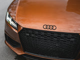 A shiny orange Audi car is parked on a concrete surface, displaying its iconic logo, sleek headlights, and a honeycomb grille. The word 