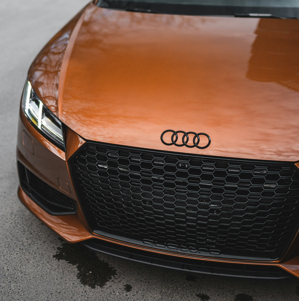 A shiny orange Audi car is parked on a concrete surface, displaying its iconic logo, sleek headlights, and a honeycomb grille. The word "quattro" is visible on the grille.