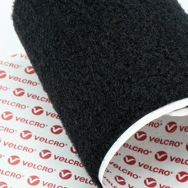 A roll of black Velcro material lies on paper printed with "VELCRO" and the company logo, demonstrating its texture and branding.