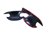 Black plastic car door handle covers lie on a white surface, featuring adhesive red tape underneath for attachment. The covers have sleek, curved designs with cutouts for handle operation.