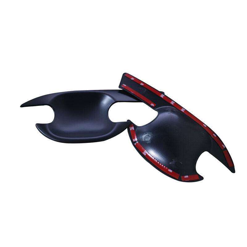 Black plastic car door handle covers lie on a white surface, featuring adhesive red tape underneath for attachment. The covers have sleek, curved designs with cutouts for handle operation.