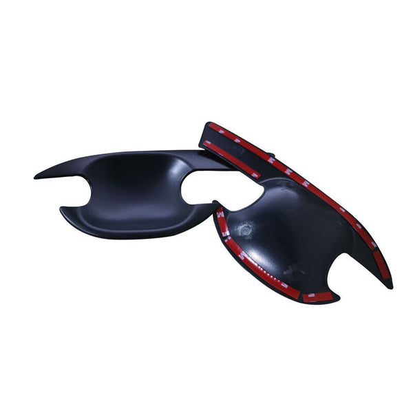 Black plastic car door handle covers lie on a white surface, featuring adhesive red tape underneath for attachment. The covers have sleek, curved designs with cutouts for handle operation.