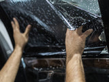 Hands apply dark film to a car window speckled with water droplets, inside a vehicle, creating a privacy tint.