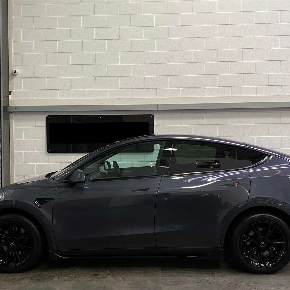 A sleek, gray car is parked indoors. It faces left, next to a metal shutter with a white brick wall in the background, creating an industrial setting.