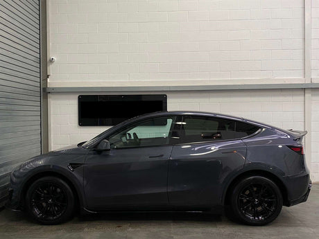 A sleek, gray car is parked indoors. It faces left, next to a metal shutter with a white brick wall in the background, creating an industrial setting.