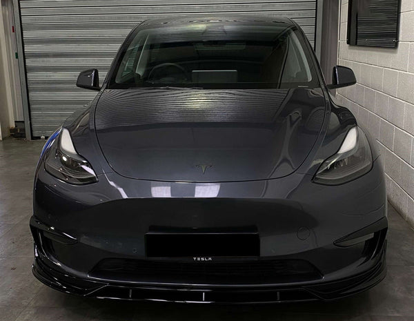 The car, a dark gray Tesla, is parked facing forward inside a garage with a closed, corrugated metal door and white brick walls. The front license plate reads "TESLA."