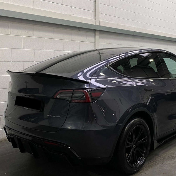 A dark-colored car with a spoiler is parked indoors against a white brick wall. Text on the car reads "DUAL MOTOR." Reflections are visible on its glossy surface.