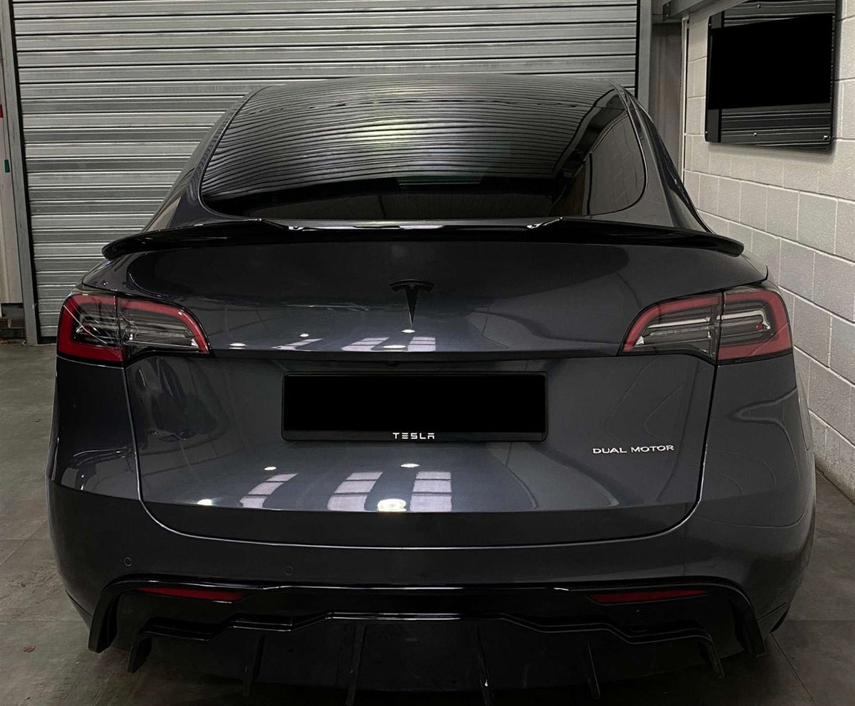 A black Tesla car is parked inside a garage. The back features "TESLA" and "DUAL MOTOR" text, with a closed metallic shutter and a wall-mounted screen in the background.