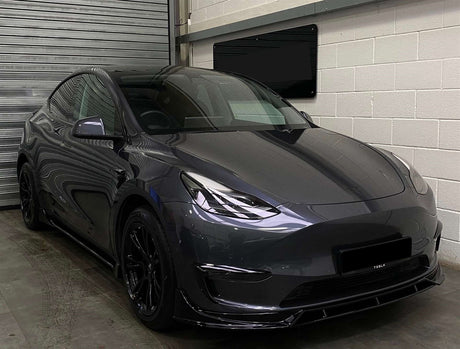 A sleek, gray car sits stationary, facing front-left, in an indoor garage with roller doors and white walls. "TESLA" is visible on the front bumper.