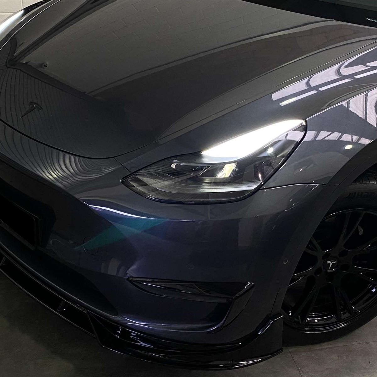 A dark-colored Tesla car with illuminated headlights is parked indoors. The focus is on the front and side, displaying its sleek design and black wheels in dim lighting.