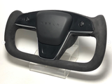 A black Tesla steering wheel with a yoke design, resting on a clear stand. The center features the Tesla logo embossed, and buttons are visible on either side.